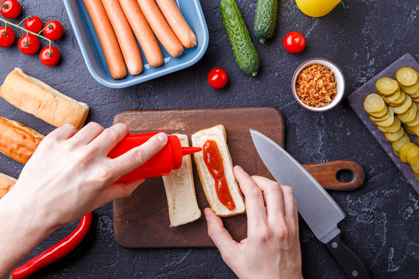 Picture on top of table with ingredients for hot dogs, cutting board, man's hands with knife - Photo, Image