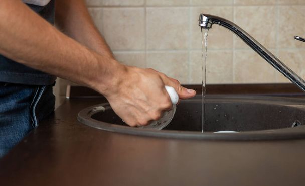 Foto de lado de las manos del hombre lavando taza transparente en fregadero en la cocina
 - Foto, imagen
