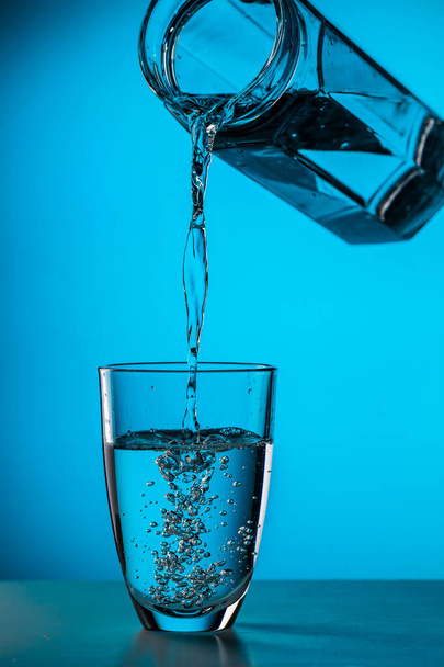 Man giet water uit glas in glas op blauwe achtergrond in studio - Foto, afbeelding