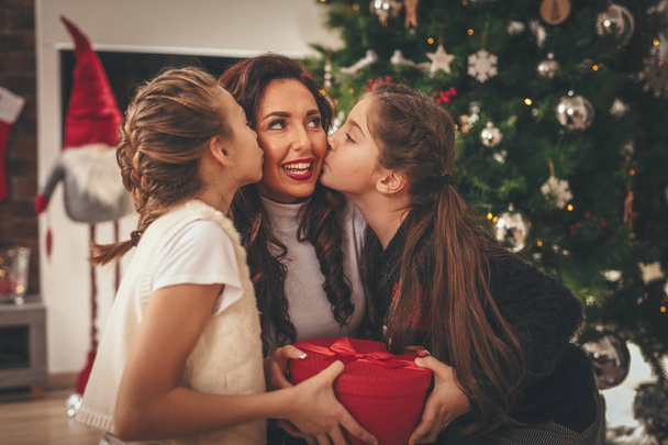 Две улыбающиеся девочки и их мама сидят у елки. Они дарят подарок в красной коробке своей матери, целуя ее
. - Фото, изображение