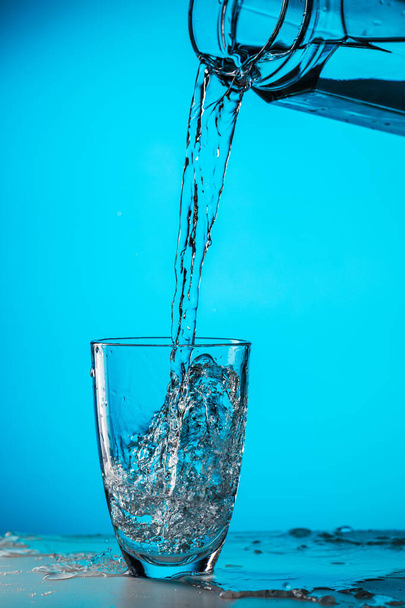 Άνθρωπος ρίχνει νερό από γυαλί σε γυαλί σε μπλε φόντο στο στούντιο - Φωτογραφία, εικόνα