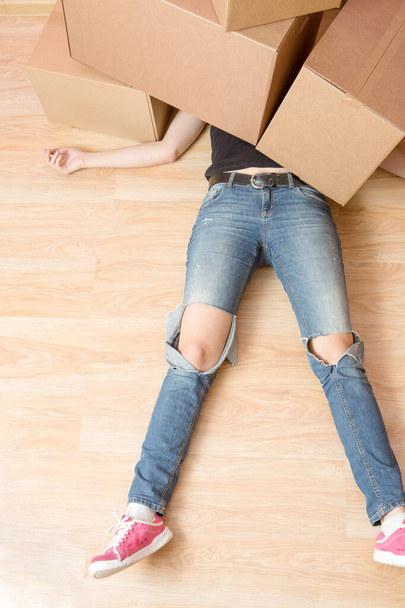 Image of woman in jeans lying under cardboard boxes in new apartment - Photo, Image