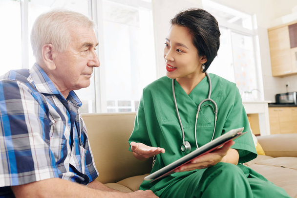 Uśmiechnięta pracownica szpitala odwiedzająca starszego pacjenta w domu, pokazująca mu informacje na tablecie i omawiająca wyniki badań medycznych - Zdjęcie, obraz