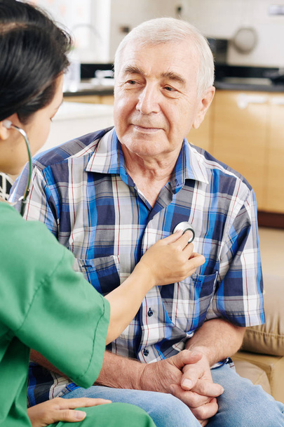 Χαμογελώντας ηλικιωμένος άνδρας που κοιτάζει τον εργαζόμενο του νοσοκομείου με πράσινη ποδιά ακούγοντας τον χτύπο της καρδιάς του όταν τον επισκέπτεται στο σπίτι - Φωτογραφία, εικόνα