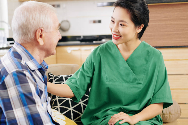 Positivo giovane asiatica caregiver femminile parlando con il paziente anziano e discutendo la sua salute e le esigenze quando visita il suo a casa sua
 - Foto, immagini