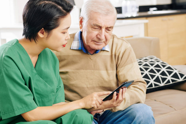 Caregiver explaining senior patient how to use application on smartphone to reach his physician - Photo, Image
