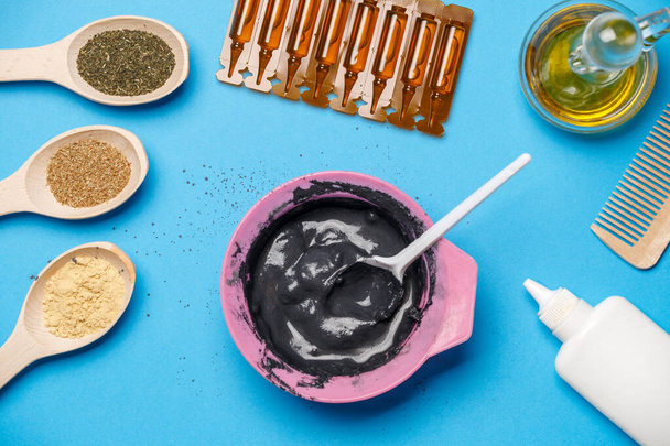 Photo from above with pink cup with black clay, mask oils, wooden spoons, combs, with spoon on blue table - Photo, Image