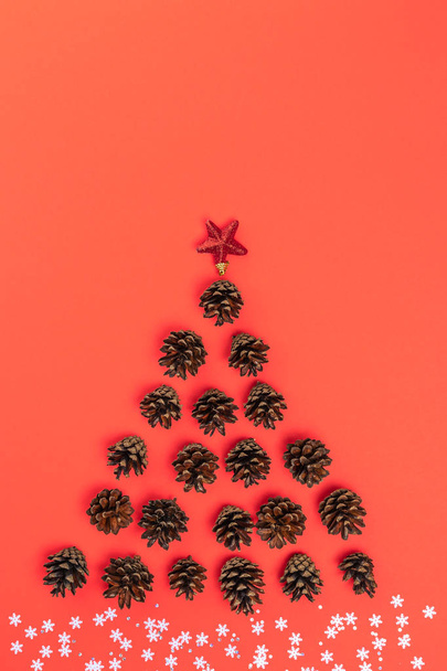 Creative layout of Christmas tree made of fir cones, star, with white snowflakes and sequins as snow on red background. Flat lay, top view. Nature New Year concept - Zdjęcie, obraz