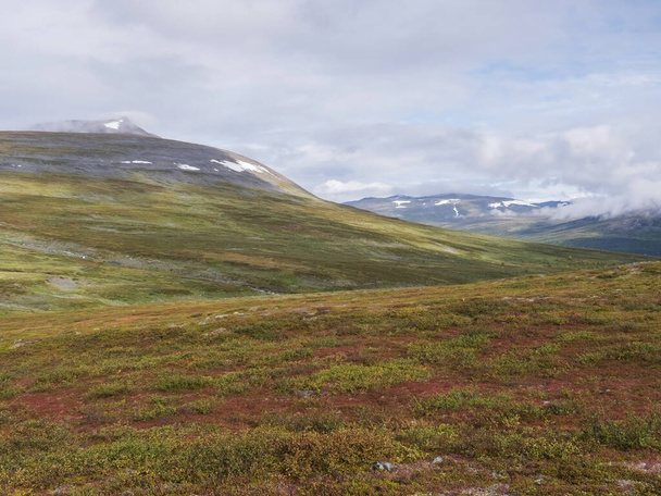 Landscape of Lapland nature at Kungsleden hiking trail with colorful mountains, rocks, autumn colored bushes, birch tree and heath in dramatic light and clouds - Fotó, kép