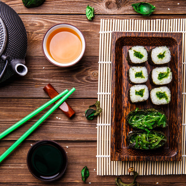 Japanilaisia rullia, vihreitä tikkuja, teekannu puupöydällä
 - Valokuva, kuva