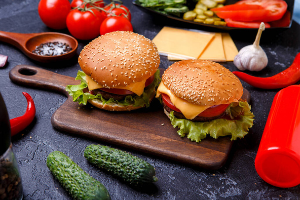 Image of two hamburgers on wooden board, cheese, tomato on black table - Фото, изображение