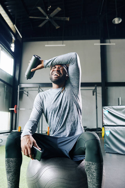 Κουρασμένος αλλά ευτυχισμένος αθλητής σκούπισμα ιδρώτα μπροστά στο μέτωπό του, όταν στηρίζεται σε μπάλα γυμναστικής μετά την άσκηση στο γυμναστήριο - Φωτογραφία, εικόνα