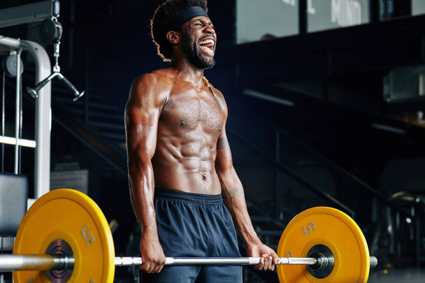hemdlos starker junger Sportler schreit beim Heben schwerer Gewichte im Fitnessstudio - Foto, Bild