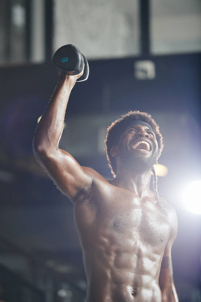 Portrait of excited shirtless muscular Black man lifting dumbbell in gym - Fotó, kép