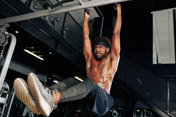 Serious concentrated fit man lifting legs when hanging on bar in gym - Photo, Image
