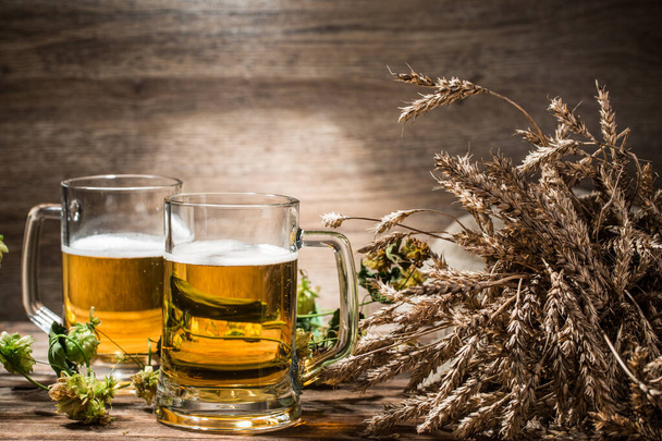 Two mugs of beer next to hops and spikelets on empty wooden background - Фото, изображение