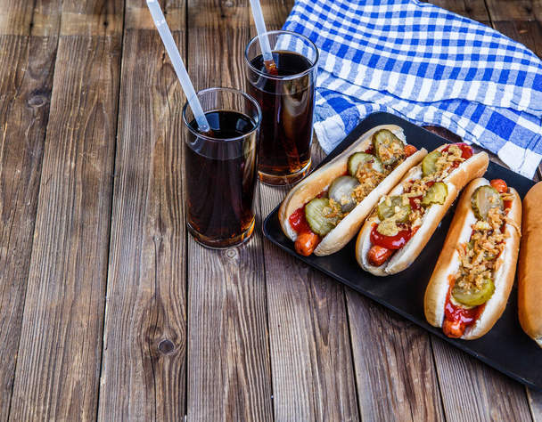 Cachorro-quente americano com picles, cebola, ketchup, mostarda e dois refrigerantes no fundo de madeira
 - Foto, Imagem