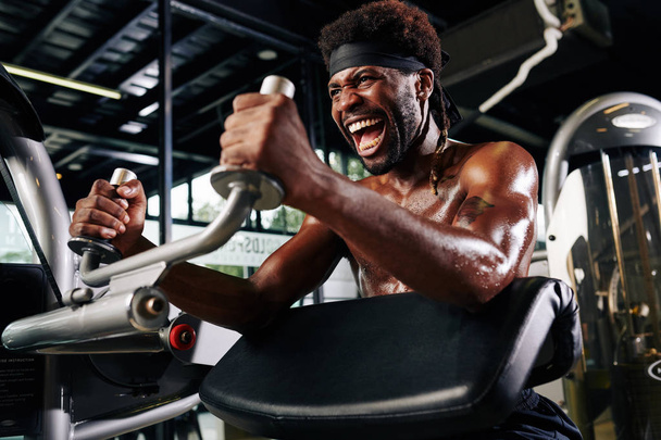 Strong young man shouting to encourage himself when doing exercise in gym machine - Photo, Image