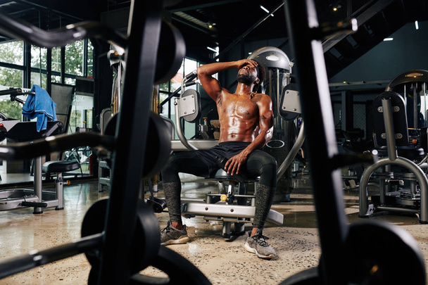 Zmęczony młody człowiek z muskularnym ciałem siedzący w siłowni i odpoczywający po treningu - Zdjęcie, obraz