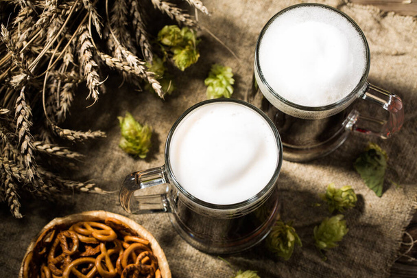 zwei Becher Bier mit Weizen und Hopfen, Brezenkorb auf Leinentuch auf Holztisch - Foto, Bild