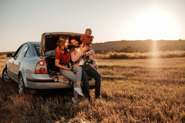 Šťastná mladá rodina maminka a tatínek s jejich malý syn těší letní víkend piknik sedí na kufru auta mimo město na poli při Sunny Day Sunset, Dovolená a Road Trip Concept - Fotografie, Obrázek