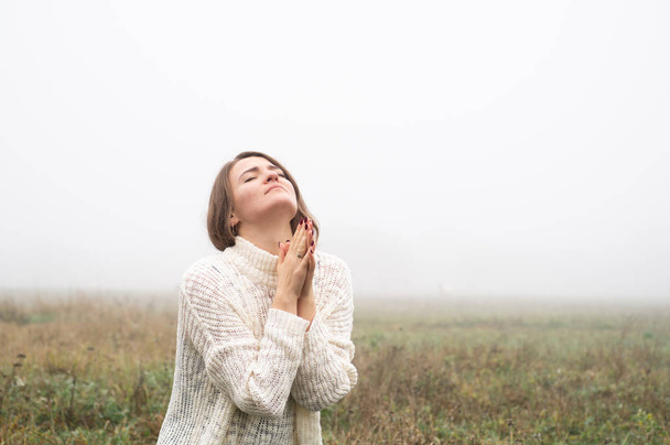 Девушка закрыла глаза на колени, молясь в поле во время прекрасного тумана
.  - Фото, изображение