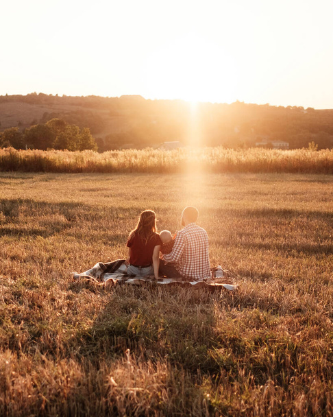 glückliche junge Familie Mama und Papa mit ihrem kleinen Sohn genießen Sommerwochenende Picknick außerhalb der Stadt auf dem Feld bei sonnigem Sonnenuntergang, Ferienzeit-Konzept - Foto, Bild