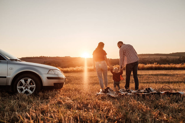 glückliche junge Familie Mama und Papa mit ihrem kleinen Sohn genießen Sommerwochenende Picknick auf dem Auto außerhalb der Stadt auf dem Feld bei sonnigem Tag Sonnenuntergang, Urlaub und Roadtrip-Konzept - Foto, Bild