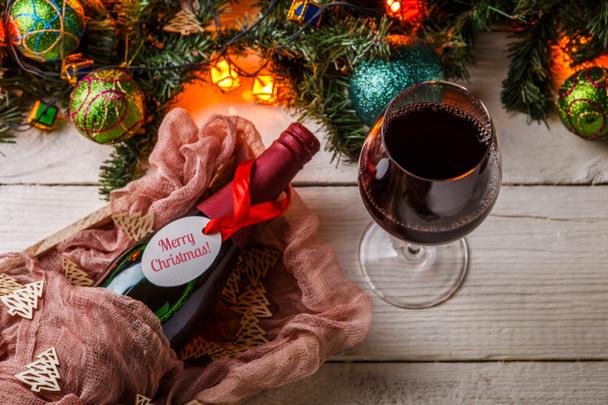 Изображение еловых ветвей, новогодние балы, горящая гирлянда, бутылка вина и бокал на белом столе
 - Фото, изображение