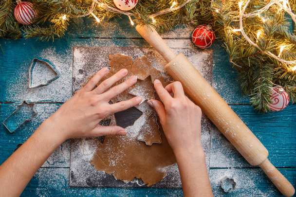 Photo de mains humaines, rouleau à pâtisserie, branches d'épinette, guirlandes, moules à biscuits sur table bleue
 - Photo, image