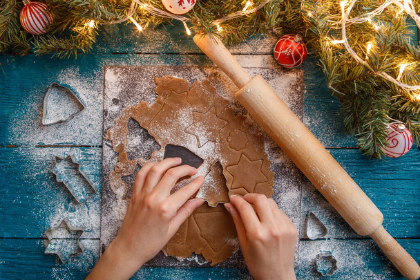 Photo de mains humaines, rouleau à pâtisserie, branches d'épinette, guirlandes, moules à biscuits sur table bleue
 - Photo, image