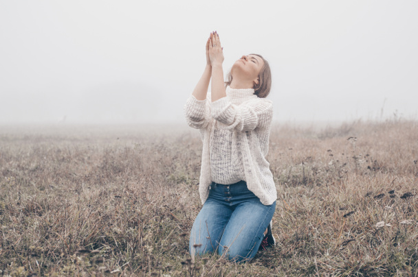 Dziewczyna zamknęła oczy na kolanach, modląc się na polu podczas pięknej mgły.  - Zdjęcie, obraz