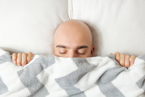ein junger Mann liegt mit geschlossenen Augen im Bett unter einer Decke und hat es morgens schwer aufzuwachen. - Foto, Bild