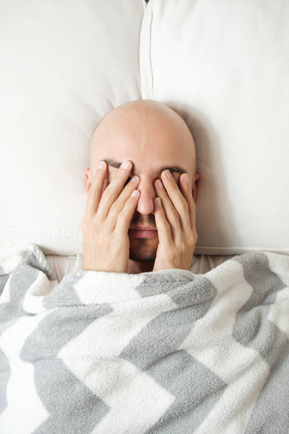 ein junger Mann liegt mit geschlossenen Augen im Bett unter einer Decke und hat es morgens schwer aufzuwachen. - Foto, Bild