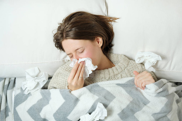 A young woman lies in bed under a blanket and blows her nose in a napkin. - Photo, Image