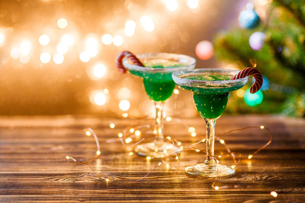 Yeni yılın bulanık arka planında yeşil kokteyl, karamel çubukları ve çelenk olan iki şarap kadehinin Noel resmi. - Fotoğraf, Görsel