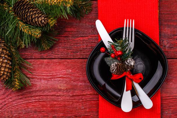 Χριστουγεννιάτικο τραπέζι θέση ρύθμιση με κόκκινη πετσέτα, μαύρο πιάτο, λευκό πιρούνι και μαχαίρι, διακοσμημένο κλαδί γκι γκι και χριστουγεννιάτικο πεύκο κλαδιά. Χριστούγεννα διακοπές φόντο. - Φωτογραφία, εικόνα