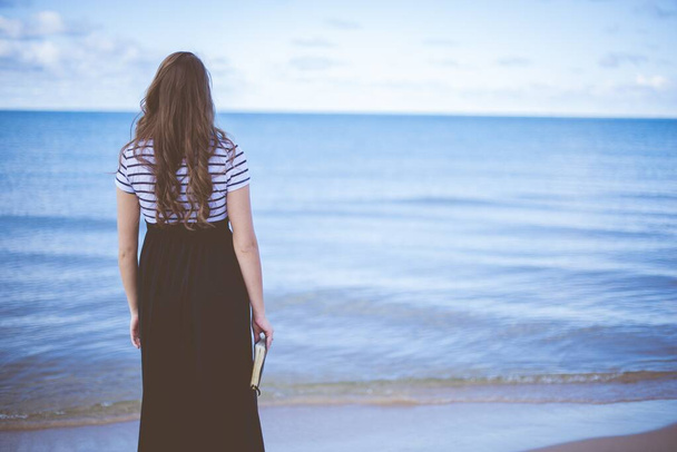 Female standing on beach shore while holding the bible with blurred background shot from behind - Photo, Image