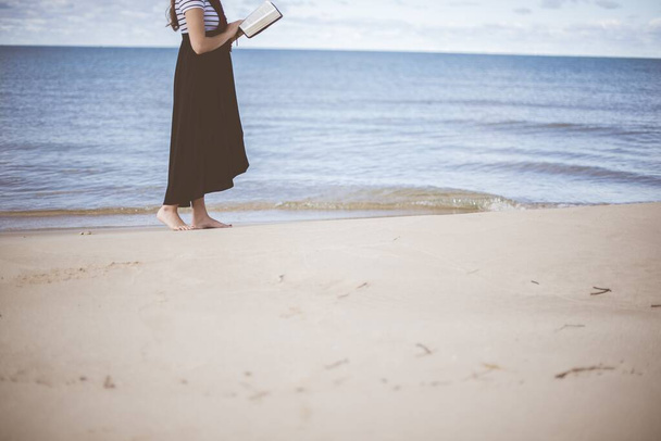 Female standing on the beach shore near the water bare feet while reading the bible - Photo, Image