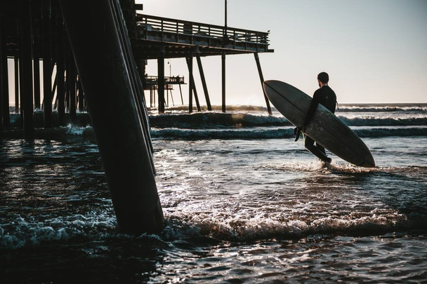 A surfboarder carrying a surfboard in the wavy ocean near the wooden pier during sunset - Fotografie, Obrázek