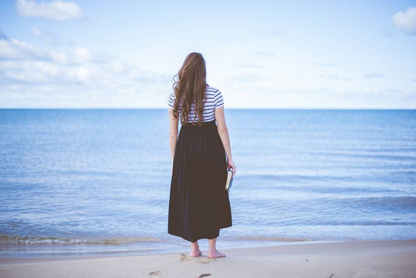 Female standing on a beach shore bare feet while holding the bible with a blurred background - Photo, Image