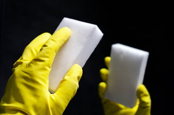 Left hand in a yellow rubber glove with a white melamine sponge in front of its reflection in the mirror in a dark room. Closeup view - Photo, Image