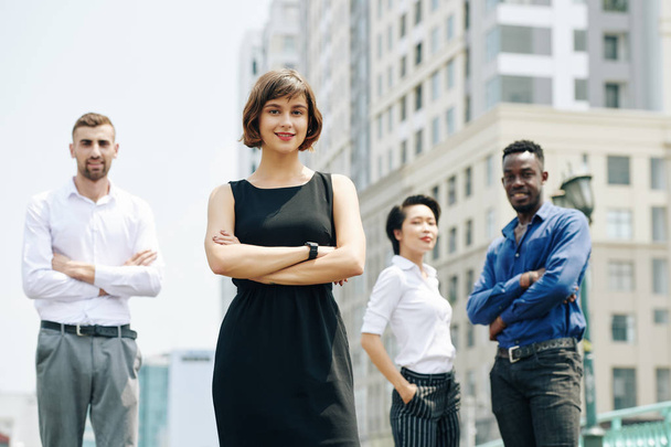 Усміхнена молода в'єтнамська бізнес-леді в чорній сукні стоїть на відкритому повітрі з колегами і дивиться на камеру - Фото, зображення
