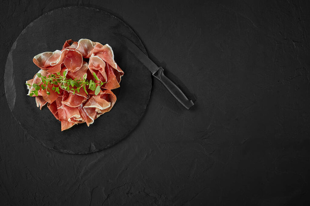 Sliced jamon with herbs and a knife are on black stone slate board against a dark grey background. Close-up shot. Top view. Copy space. - Valokuva, kuva