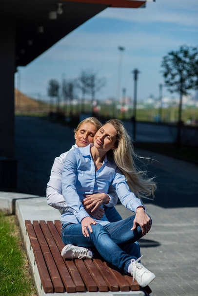 Two surprisingly similar women are sitting on a bench in an embrace and smiling. A middle-aged woman and her elderly mother. Beautiful well-groomed mother and daughter family look. - Foto, Imagem
