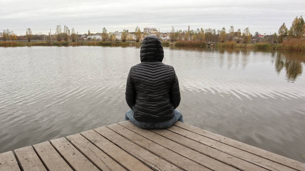 Tyttö takissa istuu puisella sillalla järven rannalla kylmällä syksyllä. Dipresia tai uneksiminen yksin. Ajattelen yksitellen luonnon kanssa
. - Valokuva, kuva