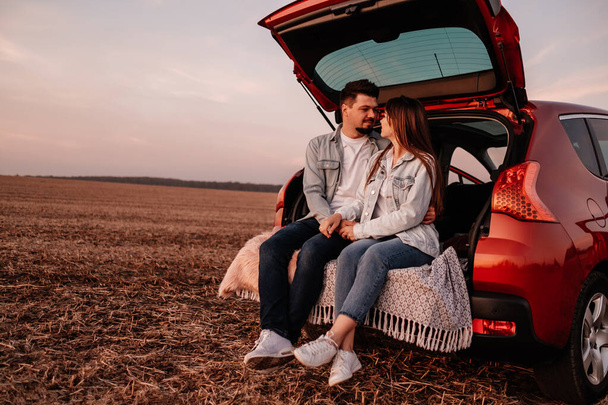 Młoda szczęśliwa para ubrana jak w białej koszuli i dżinsach siedzi na ich nowym bagażniku samochodu, piękny zachód słońca na polu, wakacje i podróży Concept - Zdjęcie, obraz