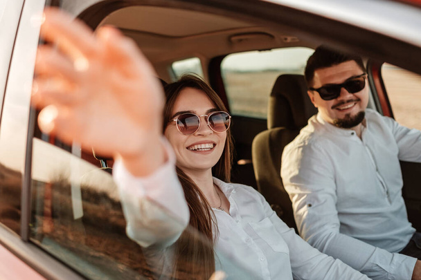 junges glückliches Paar in weißem Hemd und Jeans genießt Roadtrip am neuen Auto, schönen Sonnenuntergang auf dem Feld, Urlaub und Reisekonzept - Foto, Bild
