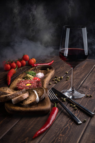 Beef Tartar with a raw egg yolk, a glass of red wine, rye bread and chilis on a wooden board with spoon and fork. Smoke background. Shot made from above on a dark rustic texture - Zdjęcie, obraz