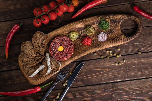 Beef Tartar with a raw egg yolk, capers, rye bread and chilis on a wooden board with spoon and fork flat lay. Shot made from above on a dark rustic texture - Zdjęcie, obraz